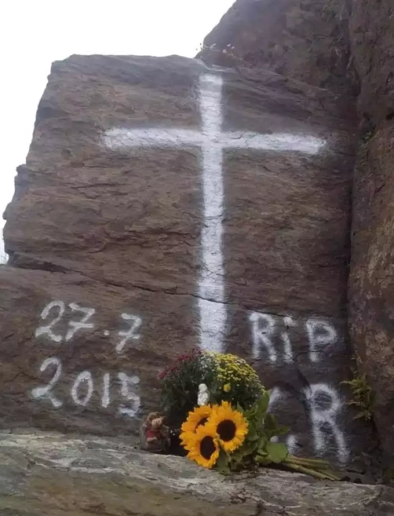 Kors og dødsdato tegnet på fjellvegg, blomster lagt ned foran.